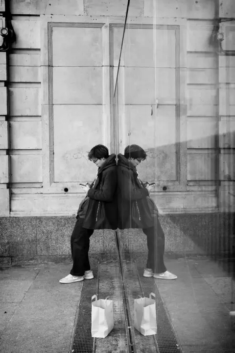 Sandy McCune A figure leans against a glass window causing a mirror image to appear. There is a stone building in the background of the picture. The person is engrossed in their phone and there is a paper bag on the ground beside them.