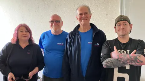 Chloe Aslett/BBC Dawn - pictured earlier - standing in a line with a bald man with glasses and a goatee, a tall man with greying hair and a smile on his face, and a younger man with tattoo sleeves and a backwards camo hat.