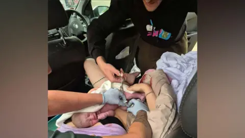 Danielle Charlton James is cutting the baby's umbilical cord. The baby is on top of the mother Sarah, who is laying in the backseat of the car, covered in various cloths. In the foreground, a paramedic who is wearing blue gloves is holding the cord steady. The baby's face is scrunched up as if he is crying.