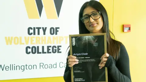 A young woman holding up a black certificate in a gilt frame, in front of a sign for City of Wolverhampton College. The woman has long black hair, dark glasses and is wearing large black-framed glasses.