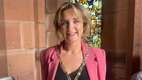 Catherine McDaid stands in front of a red brick background and a stain glass window. she is wearing a pink jacket and a black blouse. He hair is up and she is wearing a deputy mayoral chain.