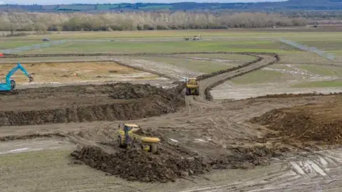 Project Universal An aerial view of the Kempston Hardwick Universal site, showing earthworks carried out by construction trucks and diggers. There is a bright blue digger on the back left, and two yellow trucks in the centre. There are trees and hills in the background.