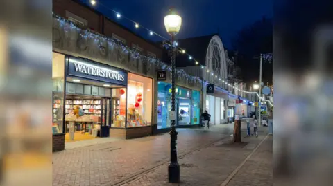The image shows a high street with lit up shops like Waterstones and Superdrug. In the centre of the image is a streetlight that has Christmas lights hanging from it. A white sheet has been stuck to the neck of the structure.
