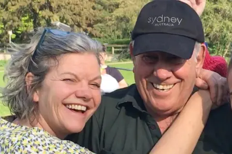 Rebecca Holder Rebecca holds her arms around her dad's shoulders. Both smile at the camera, it is a head and shoulders shot of them both. Bob wears a black cap and black polo shirt, Rebecca has chin length wavy hair, with her glasses perched on top of her head. 