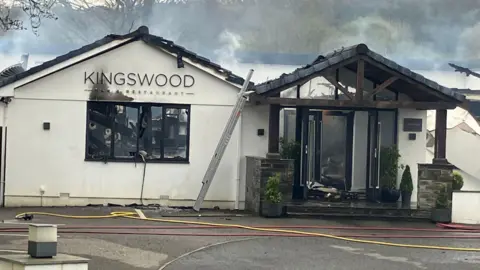 A white building which has fire damage to the front. There is text above a large window on the left-side of the building which says 'Kingswood bar and restaurant'. On the left is a porch with black double-doors. There are yellow and red wires running in front of the building. 