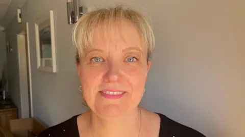 John Devine/BBC Jayne Vango is standing in a hall which has light fittings and pictures hung up. She has blonde hair in a ponytail and is wearing a black T-shirt and a silver necklace.
