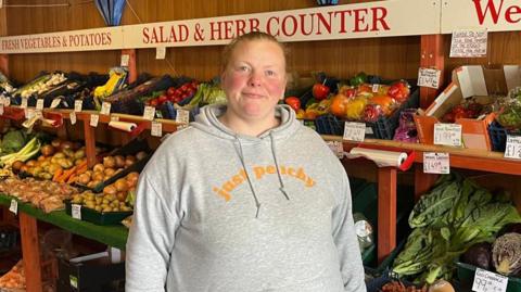 Kelly Maughan - a woman in a greengrocers wearing a grey hoodie