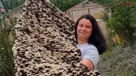 Zina A woman with long dark hair holding up a knitted rug out of brown and white wool. The woman is wearing a grey t shirt. 