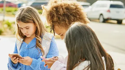 Getty Images The image shows children standing in a circle looking at their phones outside a school.
