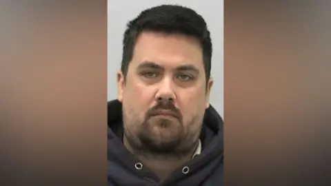 Mugshot of Scott McKenzie. He has a large head with a wispy black beard and short thick black hair.