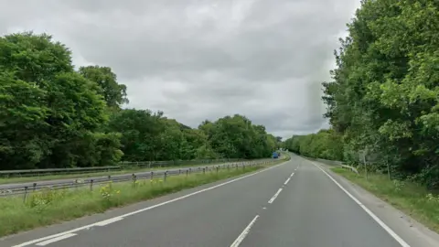 The A66 road has two lanes on either side of a metal central reservation barrier. The road is bordered by lush green trees and there is a patch of grass down the middles too. There are cars in the distance.