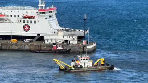 A small dredging boat is in Douglas Harbour. It has a small yellow crane-like device at one end and another piece of yellow equipment at the other. The Manxman ferry is visible on the other side of the pier.