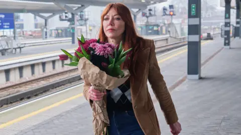 Govia Thameslink Railway Diane Morgan holds a bouquet of red and pink flowers as she walks down an empty train platform