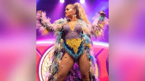Carl Sukonik Singer Rita Ora with long blond hair in a high ponytail performing on stage in 2018 wearing a multi-coloured feather coat and lilac, yellow, navy and turquoise knitted bodysuit with feathers. The stage background is purple and pink with white spotlights shining on her.