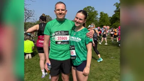 FAMILY HANDOUT An 18-year-old woman in a green Macmillan Cancer Support top and black shorts with brown hair in a ponytail standing next to her dad who is also wearing a green top and black shorts. There are several runners walking about behind them.