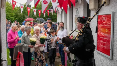 Many people, young and old, gathered outside Kilchoan Post Office to celebrate its reopening at a new location. A piper is stood outside the door in traditional dress, playing his bag pipes.