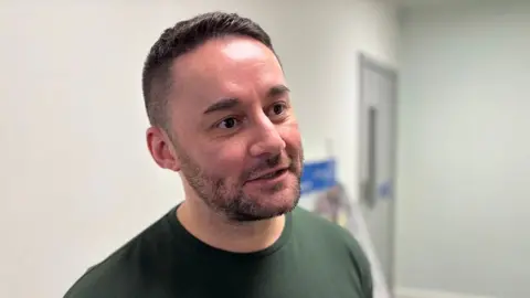 Liam Edmondson is speaking to someone who is out of shot. He has short brown hair, shaved at the sides and a trimmed beard. He is wearing a dark green t-shirt.