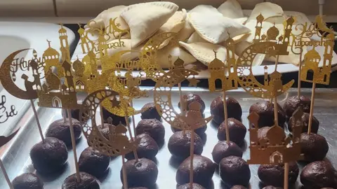 Aisha Sadiqa A tray of samosas, raw and ready to cook, piled up on a white tray. In front of those is another, metal, tray with about 28 brown cake pops laid out in rows. Stuck into these are sticks with gold cardboard or foil symbols attached to the tops. They show the outlines of mosques, crescent moons and phrases such as Happy Eid and Eid Mubarak.