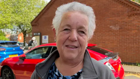 Christine Jones has grey hair and is wearing a blue shirt with a grey jacket on top. Behind her is a red car. She is smiling.