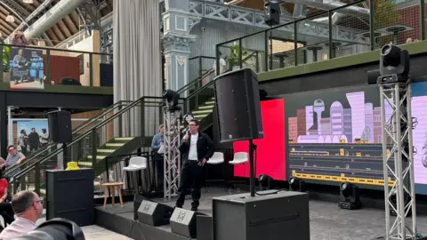 LDRS Andy Burnham speaking on stage with a screen behind him showing an underground system with sky scrapers above