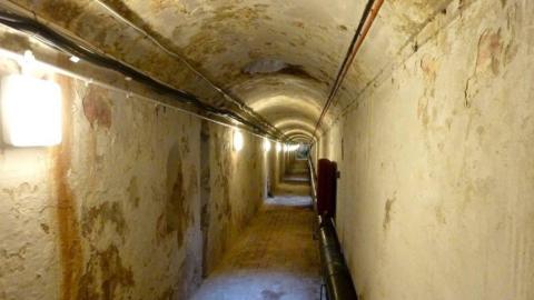 A tunnel beneath the Royal Pavilion in Brighton. The tunnel has straight side and a curved roof. The walls are white but the paint is peeling away. There is a square light on the left wall.