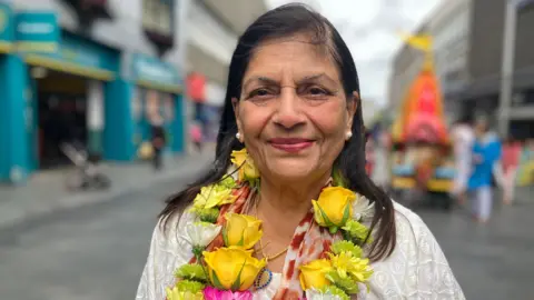 Manjula Sood stood in a city street. She has long dark hair and is wearing a necklace made of flowers.
