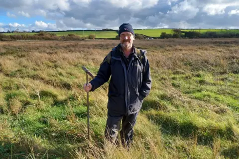 National Peatland Action Programme A person wearing outdoor clothing, a dark knitted hat and a backpack stands in a wide expanse of rough grassland, holding a long metal probe used for surveying peat or soil. Tall grasses surround the person, with shallow hollows and uneven ground visible in the foreground. Beyond the wetland, green agricultural fields and hedgerows stretch across gently rolling countryside. Large grey and white clouds dominate the sky, while bright sunlight breaks through to illuminate the vegetation, highlighting the contrast between the golden-brown grasses and the vivid green fields in the distance. The image illustrates fieldwork taking place in a peatland landscape.