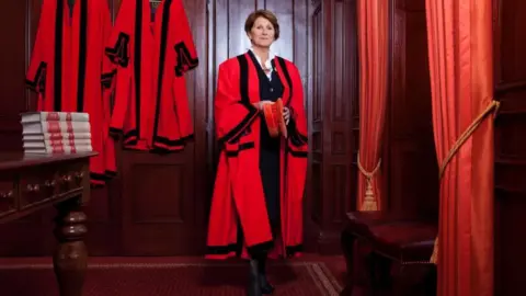 Collette Crill Collette Crill is smiling as she stands in her law chambers in her robes holding a red hat. She has a short bob and is wearing a black suit under her robes. The robes are red with black stripes and there are more hanging up on the wall behind her. To her left is a table with leather bound books on it and to her right is a leather stool and a pair of red curtains pinned open with gold coloured tie backs.
