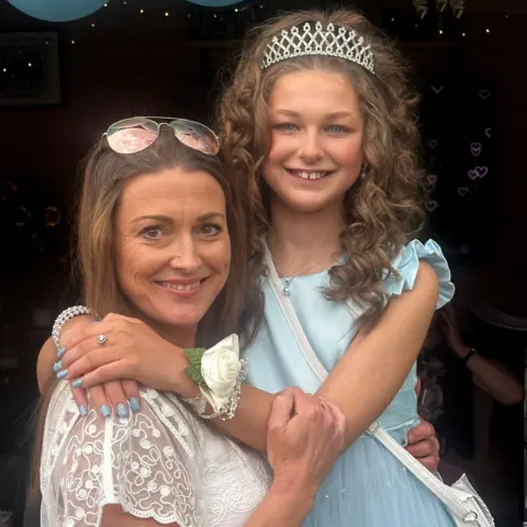 Amanda Axiak A woman and a girl with their arms around each other, smiling and looking at the camera. The woman, on the left, has long brown, straight hair and is wearing a lacey white top and sunglasses on her head. The girl has long wavy brown hair with a silver tiara in it. She is wearing a pale blue dress and white cross-body bag and has a white corsage on her wrist, a silver heart-shaped ring on one finger and pale blue nail polish.