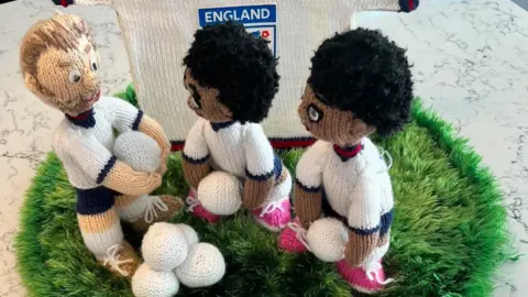 A knitted display features three figures of England players holding small white balls. There is also a pile of four white balls on the ground - a circular patch of green wool. A small knitted England shirt is standing behind them.