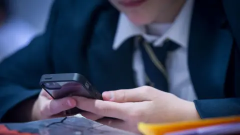 A school pupil wearing a shirt, tie and blazer holding a mobile phone in a classroom. 