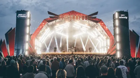 Aled Llywelyn | Eisteddfod Genedlaethol A large crowd watches a band perform on an illuminated stage.
