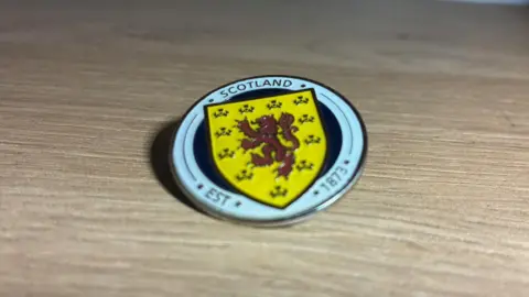 A circular metal badge with the Scotland crest showing a red lion rampant on a yellow background upon a navy circle with a white border. Text around the outside reads "Scotland Est 1873"