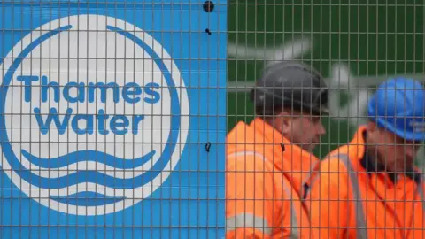 Two men in orange hi-vis jackets and safety helmets stand behind a wire fence. To their left is a blue and white Thames Water logo.