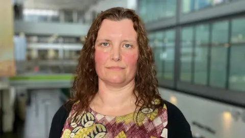 Dr Amy Doherty stands inside a large building. She has shoulder length brown wavy hair. She is wearing a purple dress with green floral markings on it and a black cardigan over it.