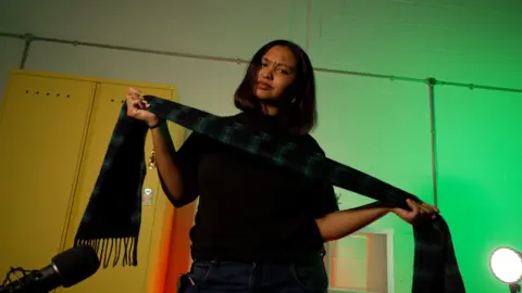 A woman with shoulder length dark hair holding a tartan scarf.