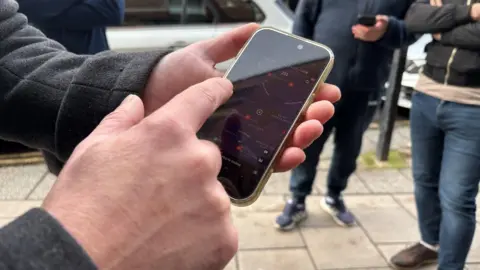 A close-up picture of a man holding a phone, showing an Uber map. He is holding the phone with one hand and pointing at it with the other. In the background there are people standing on the pavement. 