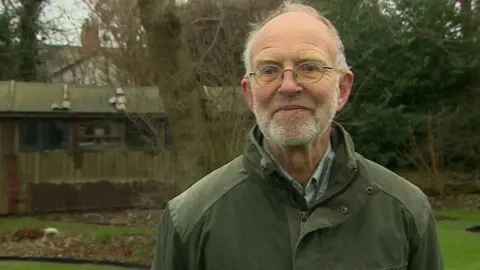 Stephen Reeves is standing in his garden. He is wearing a green jacket and is an elderly man with white stubble and glasses.