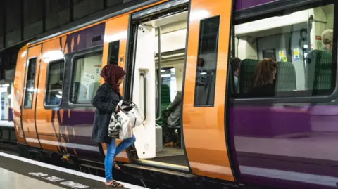 A woman wearing a red hijab, blue coat and blue jeans is walking onto an orange and purple train carriage while holding bags.