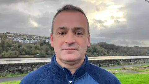 Malachy McCarron is wearing a blue fleece and has short brown hair. He is standing near a waterway, and a number of homes are visible in the background on the top of a hill.
