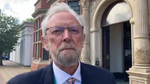 BBC/Amanda White Andy has grey hair and a beard and is standing outside a town hall building. He is wearing black-framed specs on his face, a navy blazer over a pale blue shirt and multicoloured tie.