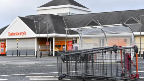 Pacemaker Trolleys and a trolley shelter in Sainsbury's car park blown over by the wind. The building is behind with orange Sainsbury's logo. There are no cars or people.