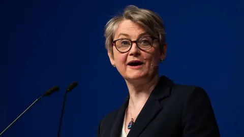 Getty Images Yvette Cooper stood in front of a lectern making a speech in front of a plain navy blue background. She is wearing a black jacket, necklace, and a white top.