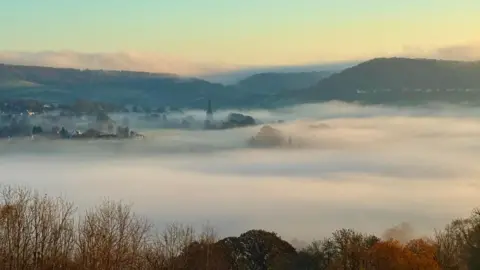 BBC Weather Watchers/Space Walker A misty, golden morning scene over valleys. A church spire and browning trees can be sticking out of the layer of cloud.