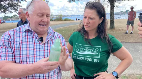 Richard Westacott-Jones is wearing a red and blue checked shirt and holding a pale green bottle. Mallory Haas in a green T-shirt with the words The Barn Pool Dig watches. They are outside in a park with the sea in the background.