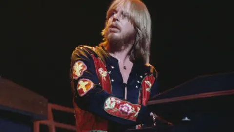 Getty Images Rick Wakeman performs on a keyboard during a concert in the 1970s. He has blond hair that reaches his shoulders and a fringe. He is wearing a black and red top.