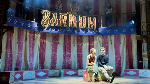 A man and a woman sat on a stage with red and yellow curtains and the writing BARNUM above it. 