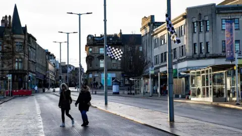SNS A view of a town's high street, with various people walking around, a bus shelter and various shops and flats. Black and white flags hang from lamp posts, showing support for the local football team St Mirren.