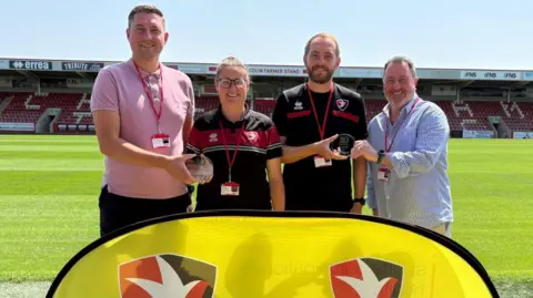 Cheltenham Town Community Trust People smiling into the camera with trophies in hand smiling. There's a large yellow banner in front of them with branding on it for Cheltenham Town FC.