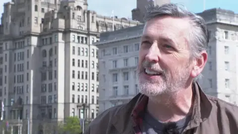 Tony Schumacher with grey hair and beard wearing a brown jacket, red and black checked shirt and grey t-shirt stands in front of the Royal Liver building in Liverpool on a sunny day.